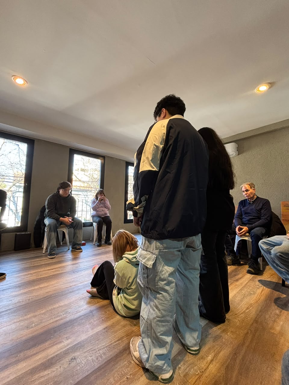 Taller en círculo: personas sentadas alrededor de la sala miran con recogimiento a dos representantes de pie y a una joven sentada en el piso.