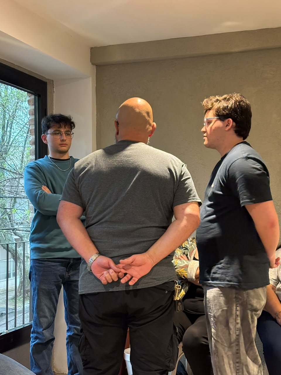 Tres representantes de pie en el centro de la sala: un hombre de espaldas, con las manos atrás, y dos jóvenes que lo miran, junto a una ventana con árboles.